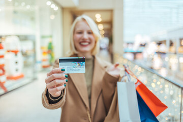 A happy beautiful blonde woman is smiling at her credit card, holding shopping bags, copying the space on the background of the shopping center. 