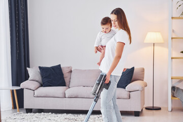 Woman uses vacuum cleaner. Mother with her little daughter is indoors at home together