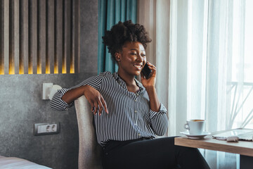 A happy business woman is talking on a mobile phone while sitting at the table of a hotel room with...