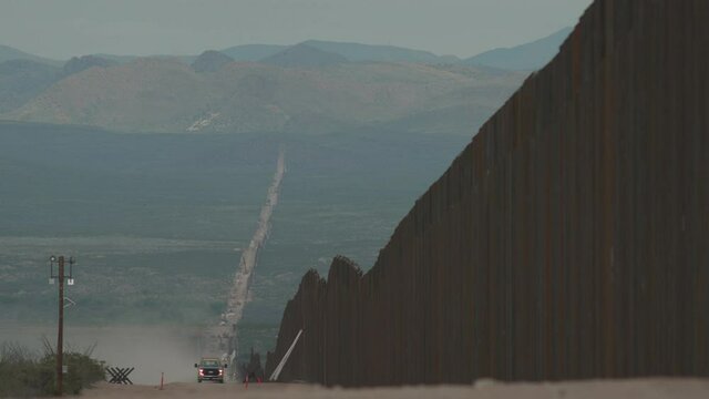 Two construction trucks with flashing lights drive towards camera alongside the newly constructed border wall between US and Mexico.