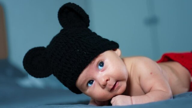 Lovely Sweet Baby In Mickey Mouse Hat Rises Head While Lying On The Bed. Beautiful Blue-eyed Child Looks Up And Opens Tiny Mouth. Close Up.