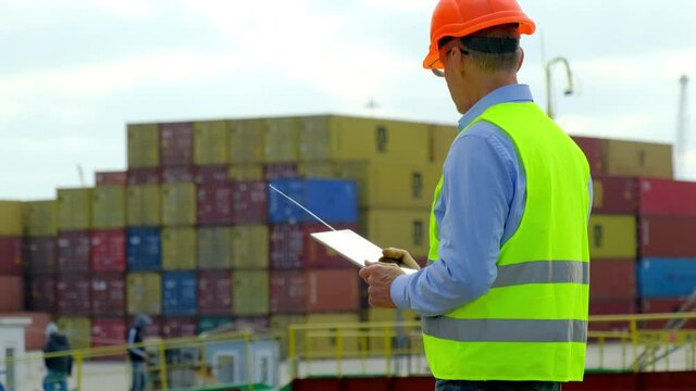 Marine port freight ship loading. Skilled engineer man in colorful with tablet computer controls employees working with metal containers in dock