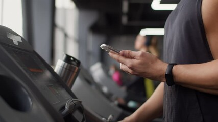 White man in the gym turns on the treadmill and walks along it, holding smartphone in his hand. man with a smart watch on his arm - Powered by Adobe