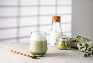 Green matcha latte in a glass on a gray background with morning shadows from the window, a bottle of milk and tea powder The concept of a vegan drink, superfoods and healthy   food. Copy space