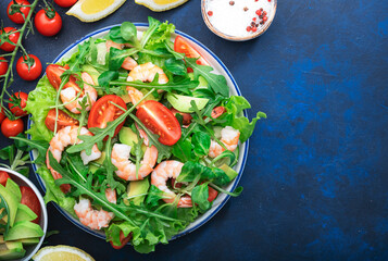 Fresh spring shrimp salad with tomatoes, arugula, avocado, and olive oil lemon dressing blue table background. Top view, copy space