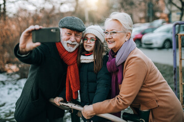 Happy senior husband and wife taking selfiephoto with their cute granddaughter in city park.