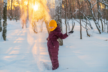 a girl in a winter jumpsuit in the forest in winter throws snow in the rays of the setting sun