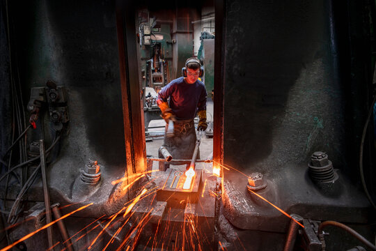 Apprentice engineer forging steel parts in hammer press in industrial forge