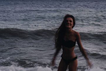 happy girl in a swimsuit enjoys freedom on beach by sea with waves. Photo with a blur from a long exposure to give an authentic atmosphere