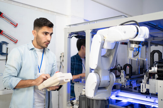 Young Caucasian male industrial engineer in a blue shirt standing using a robot controller checking and testing the work of an automation robotic arm machine before being used in an industrial factory