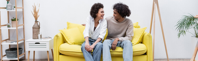 Positive african american couple looking at each other on couch at home, banner.