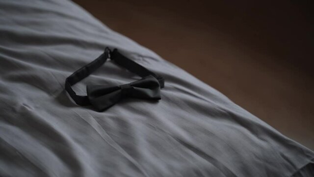Black unbuttoned bow tie lies on white crumpled bed linen. Family quarrel. Barefoot woman in veil walks past the bed. Wedding preparations.