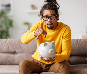 money problems. Poor upset african american man putting coin in ceramic piggy bank