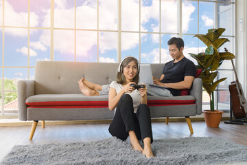 Lovely Asian couple in living room at home. young woman with headphone sit on floor playing video game, man lying on couch working with laptop computer in background at home. Love, happiness