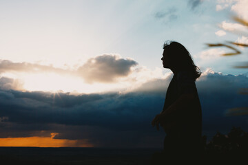 Fototapeta premium The silhouette of a young woman stands on the rocks at the top of the mountain and looks at the sunset.
