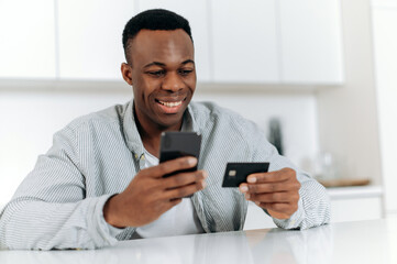 Online shopping. Satisfied black guy makes purchases from home in online stores and applications, uses smartphone and bank card to pay for purchases and delivery, enters data, looks at the card, smile