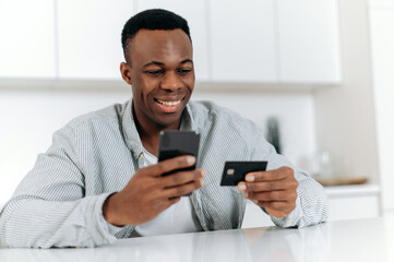 Obraz na płótnie Canvas Online shopping. Satisfied black guy makes purchases from home in online stores and applications, uses smartphone and bank card to pay for purchases and delivery, enters data, looks at the card, smile