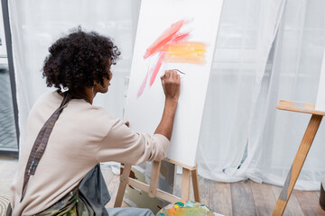 Young african american man in apron painting on canvas at home.