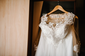 Beautiful classic lace silk wedding dress hanging on hanger in wooden room. morning preparation...