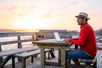 digital nomad sitting outdoors on the table on the beach with a laptop alone doing telecommuting at sunset