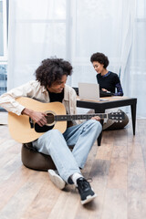 Smiling african american woman playing synthesizer near laptop and boyfriend with acoustic guitar in living room.