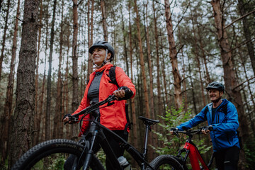 Fototapeta na wymiar Low angle view of senior couple bikers walking and pushing e-bikes outdoors in forest in autumn day.