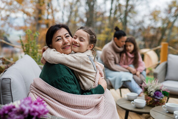 Happy little girl with grandmother sitting together wrapped in blanket outdoors in patio in autumn