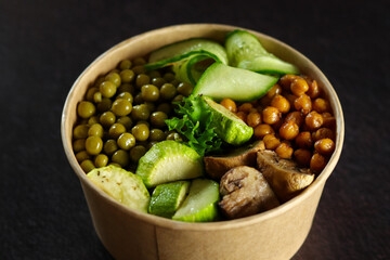 Vegetarian healthy food with chickpeas, cucumbers, zucchini, mushrooms and green peas in a paper eco container isolated on a black background. Healthy food delivery concept.
