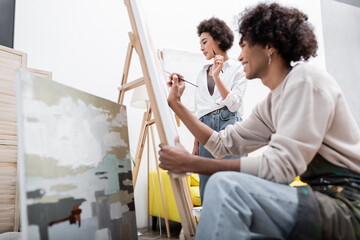 Fototapeta na wymiar Young african american woman holding paintbrush near canvas and blurred boyfriend painting at home.