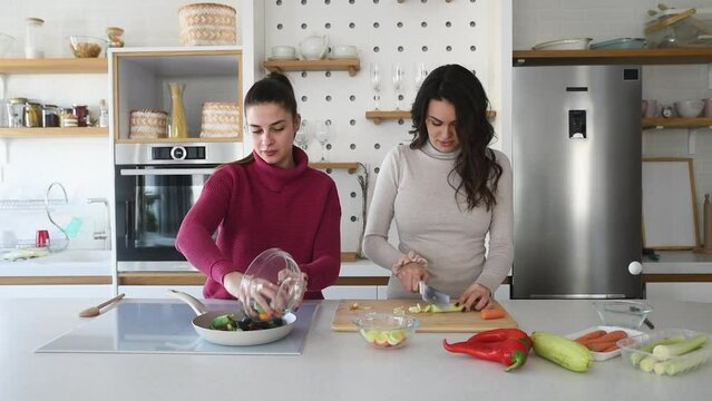 Two young happy college student roommates or business women cooking food together at their apartment. Females having fun together preparing meal in their modern kitchen at cozy home.