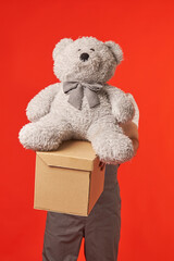 Man holds a large gray teddy bear and a cardboard box, on a red background. The courier brought a birthday present.