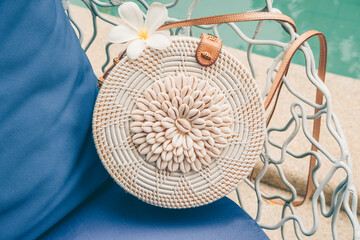 Trendy round crossbody straw bag with seashells and flower on the beautiful lounge chair near a pool. Summer outfit, vacation, holiday concept. Travel background with place for text