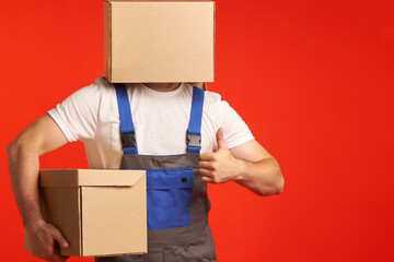 Male loader in uniform put one box on his head, and holds the other with his hand. Isolated on a red background, close-up. Shows the ok gesture with one hand. - obrazy, fototapety, plakaty