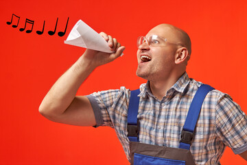 Bald man in work clothes and protective glasses sings into a rolled-up pipe made of paper, from...