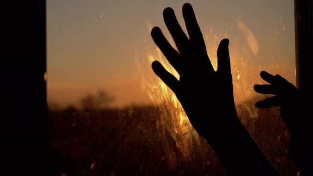 Female Hands Stretching for the Sun by the Window, Touching Dirty Glass at Home. Dawn. Early morning. Silhouette fingers hands slide over unwashed glass. Concept of suffering, hope, coronavirus, save.