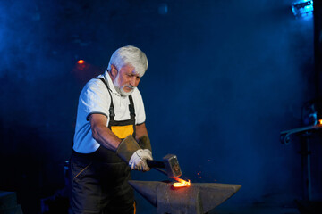 Mature caucasian craftsman in safety gloves using industrial hammer for hitting heated steel at forge. Shaping of steel on anvil. Concept of blacksmithing.