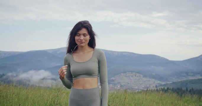 Front view of young woman with slender body walking among green grass with mountains on background. Fitness lady in sport clothes resting after outdoors activity. 