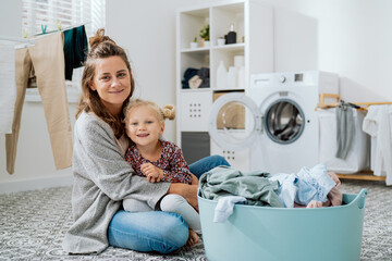 A mom sits on laundry room floor with her young daughter, a cute little girl with blonde hair tied...