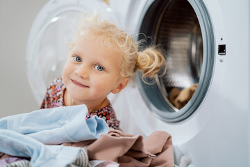 Portrait of blue-eyed girl with blonde hair tied in two buns child plays in pile of colored clothes...