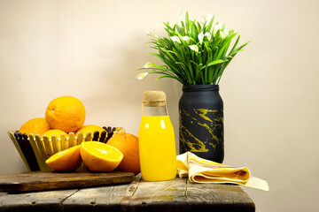 glass bottle of fresh orange juice with fresh fruits in basket and vase of lilies flowers on wooden planks table