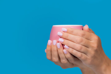 Close-up view stock photography of two female hands of european woman holding one pink mug isolated on blue background. Fingernails painted in cute pastel pink color with small painted white hearts