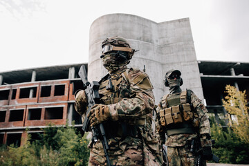 Two soldiers in camouflage uniforms with rifles in front of a building