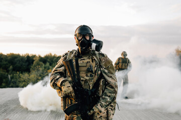 Military man in a gas mask shrouded in a smoke screen, standing with a rifle with a partner