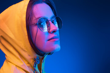 Neon close up portrait of young woman round sunglasses yellow raincoat on dark blue background