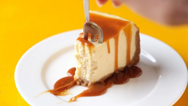 Cheesecake with caramel sauce on plate, yellow background. Taking bite of cheesecake with fork