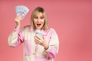 Happy winner! Image of an excited young lady, isolated over pink background. Holding a mobile phone and money.