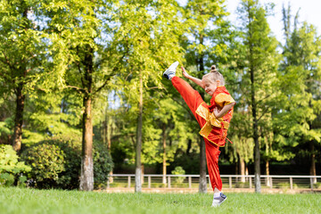 Cute little caucasian girl seven years old in red sport wushu uniform exercising in park at summer...