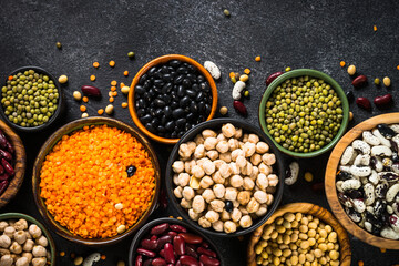 Legumes, lentils, chikpea and beans assortment in different bowls on black stone table. Top view.
