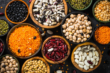 Legumes, lentils, chikpea and beans assortment in different bowls on black stone table. Top view.