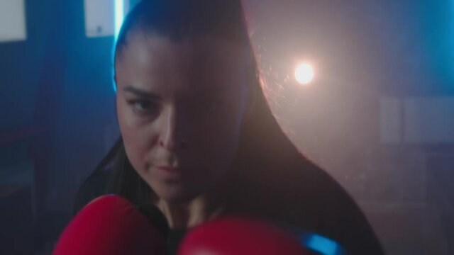 Close-up Point of view of a young brunette woman punches and kicks, looking into camera. Beautiful strong female kickboxer training at dark gym. Slow motion. High quality 4k footage